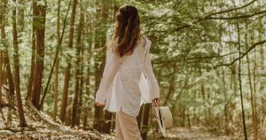 A woman is walking among trees with her back turned o the camera. She is wearing a white linen shirt, khaki pants, and is carrying a wide brimmed hat.